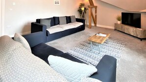 A loft-style living room with exposed wooden beams, a black sectional sofa, a wooden coffee table, and a large flat-screen TV. - Town Hall Lofts Flat 2 Settle (Settle)