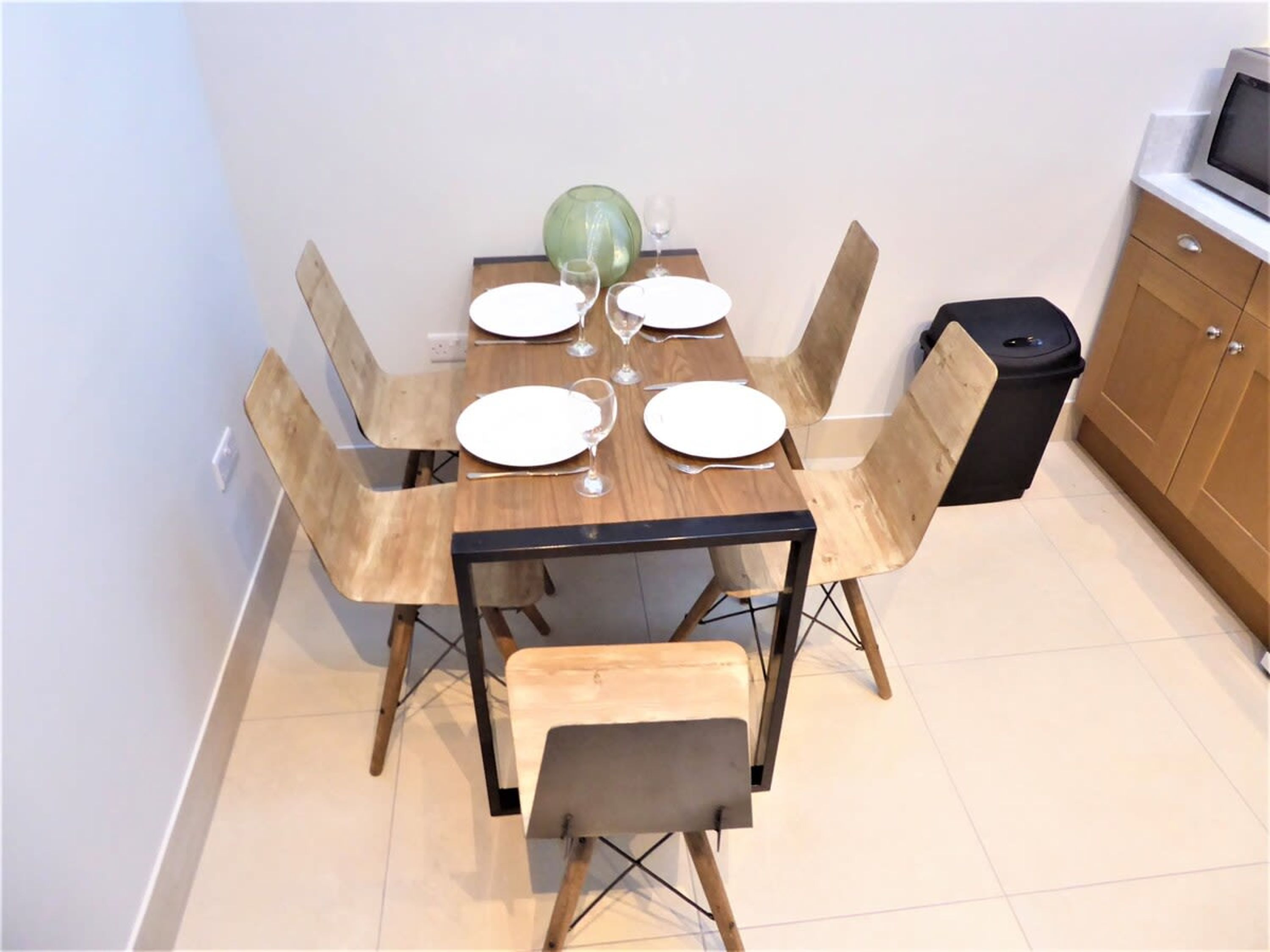 A dining area with a wooden table set for four, surrounded by chairs with wooden frames and cushioned seats. A trash bin is placed to the right of the table.