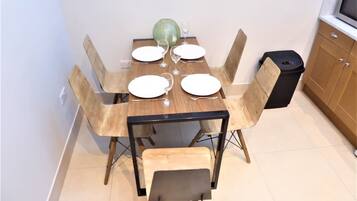 A dining area with a wooden table set for four, surrounded by chairs with wooden frames and cushioned seats. A trash bin is placed to the right of the table.