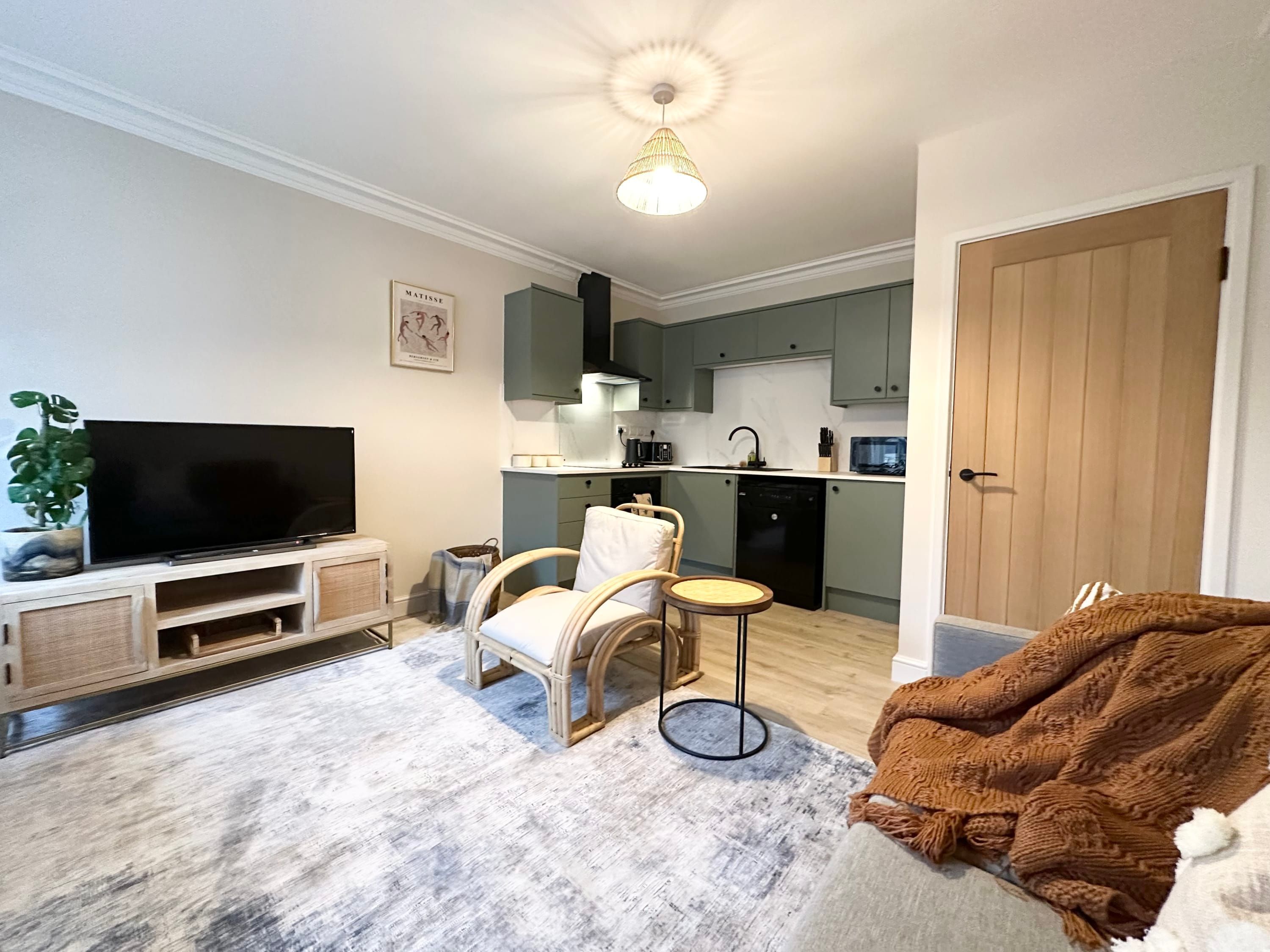 A living room with a TV, a sofa, a chair, a small table, and a kitchen in the background.