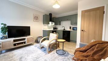 A living room with a TV, a sofa, a chair, a small table, and a kitchen in the background.