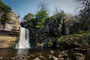 A waterfall cascading into a clear pool surrounded by rocky cliffs and sparse vegetation.