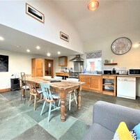 A spacious kitchen with wooden cabinets, a dining table, and chairs. There is a clock on the wall and a microwave on the counter.