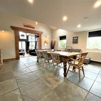 A spacious dining area with a large wooden table and six chairs, a stone-tiled floor, and a view of the outdoors through a window.