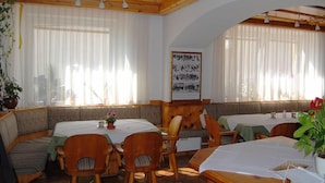 Un restaurant chaleureux, avec des meubles en bois, de grandes fenêtres ornées de rideaux légers et une œuvre d’art encadrée accrochée au mur.