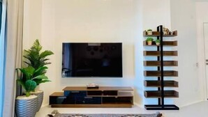 A modern living room with a flat-screen TV, a wooden entertainment unit, and a black shelving unit with plants. - Ground Spacious 1-BR Apt @ SwanLake - El Gouna (Red Sea Governorate)