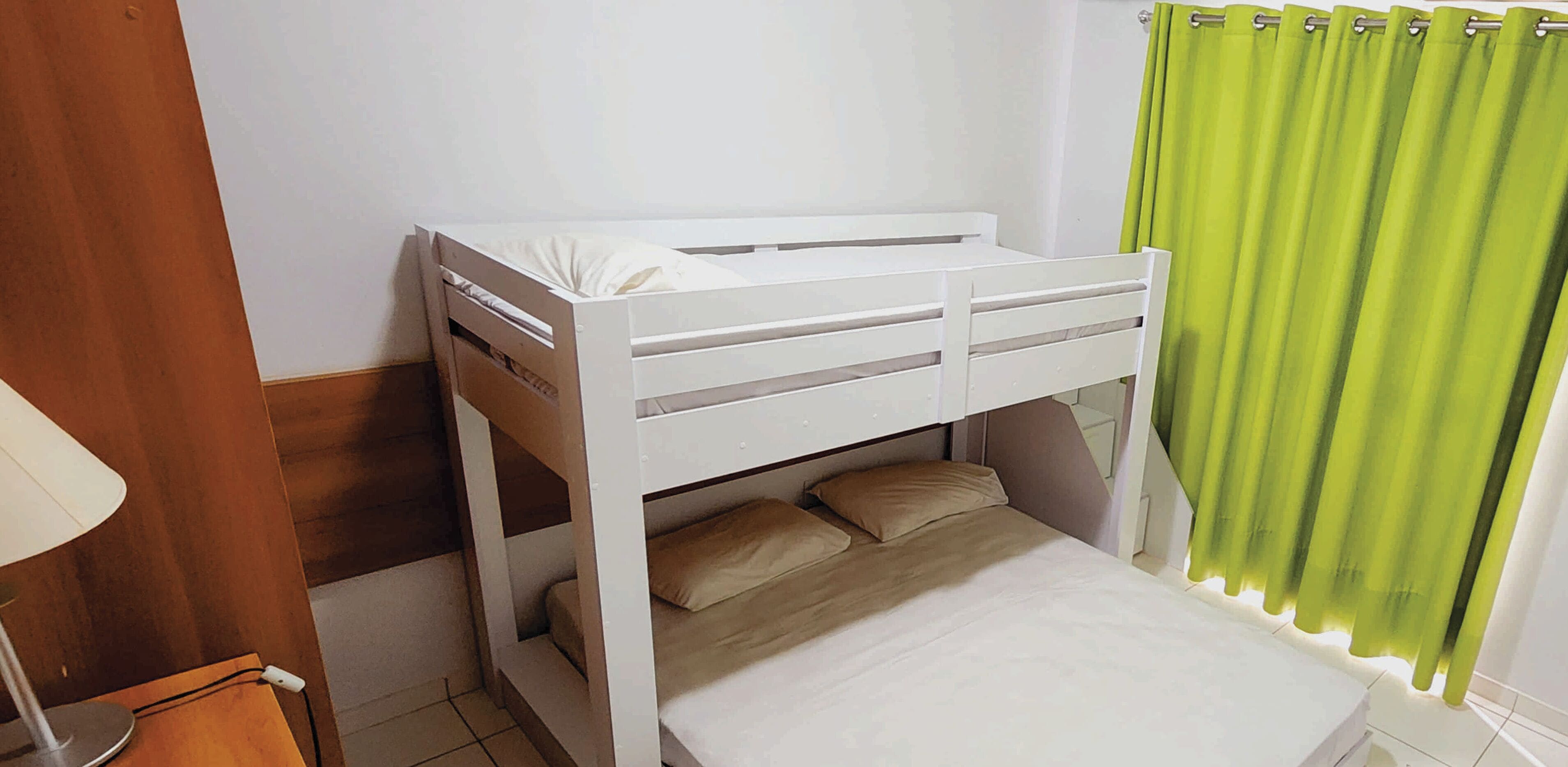 Un lit superposé avec une structure blanche et une literie beige, une table de chevet en bois avec une lampe, et un rideau vert.