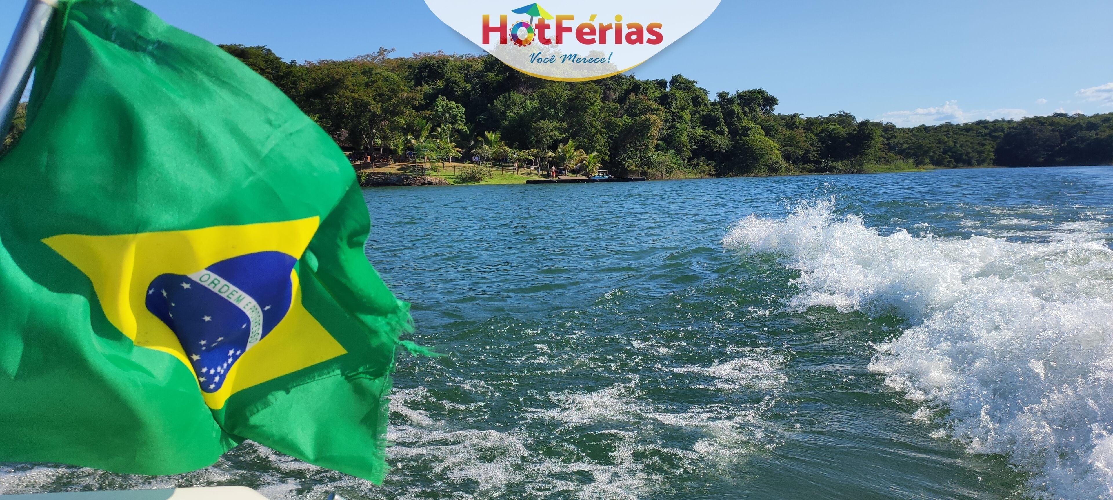 A flag waving in the wind, a body of water, and a forested shoreline.