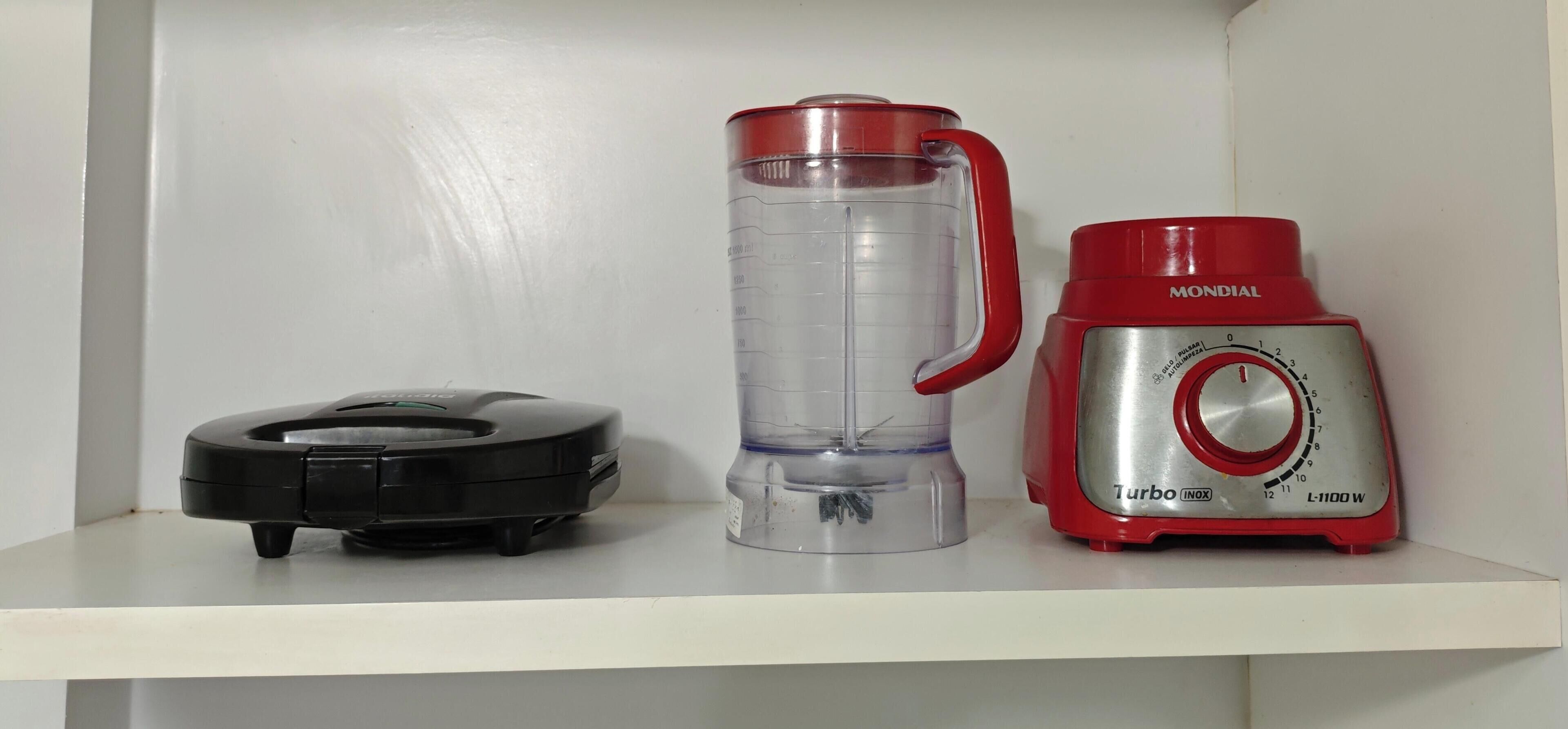 A kitchen shelf with a black circular object, a clear cylindrical container, and a red and silver blender.