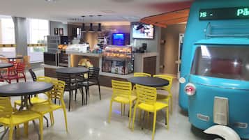 Un restaurante con sillas y mesas amarillas, una decoración con forma de autobús azul y un letrero digital que muestra