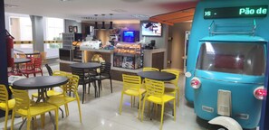 Een restaurant met gele stoelen en tafels, een blauwe decoratie in de vorm van een bus en een digitaal bord waarop "Pão de" staat.