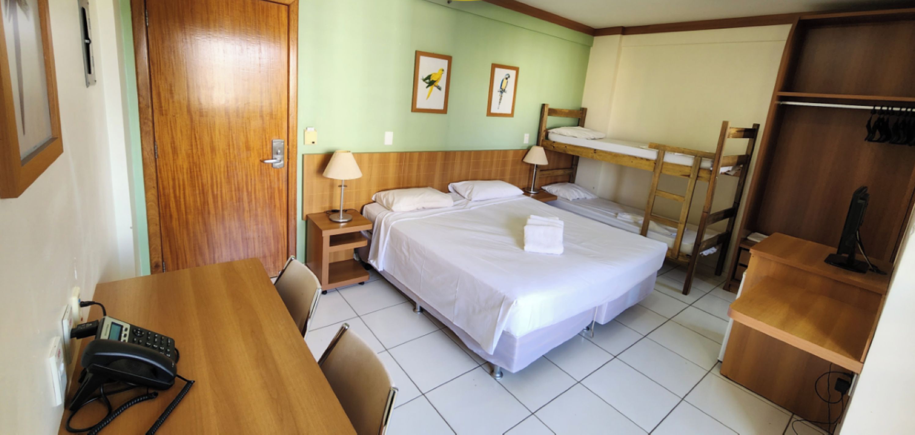 Quarto de hotel com uma cama de solteiro, uma escrivaninha com telefone, uma cadeira, uma mesa de cabeceira e uma cama beliche.