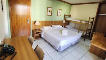 Quarto de hotel com uma cama de solteiro, uma escrivaninha com telefone, uma cadeira, uma mesa de cabeceira e uma cama beliche.