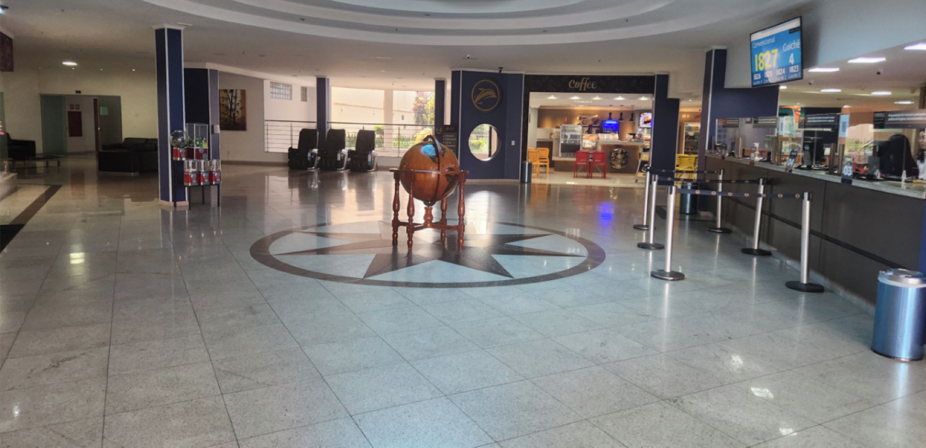 Um hall amplo com uma escultura central de um globo, uma cafeteria e um balcão de recepção.
