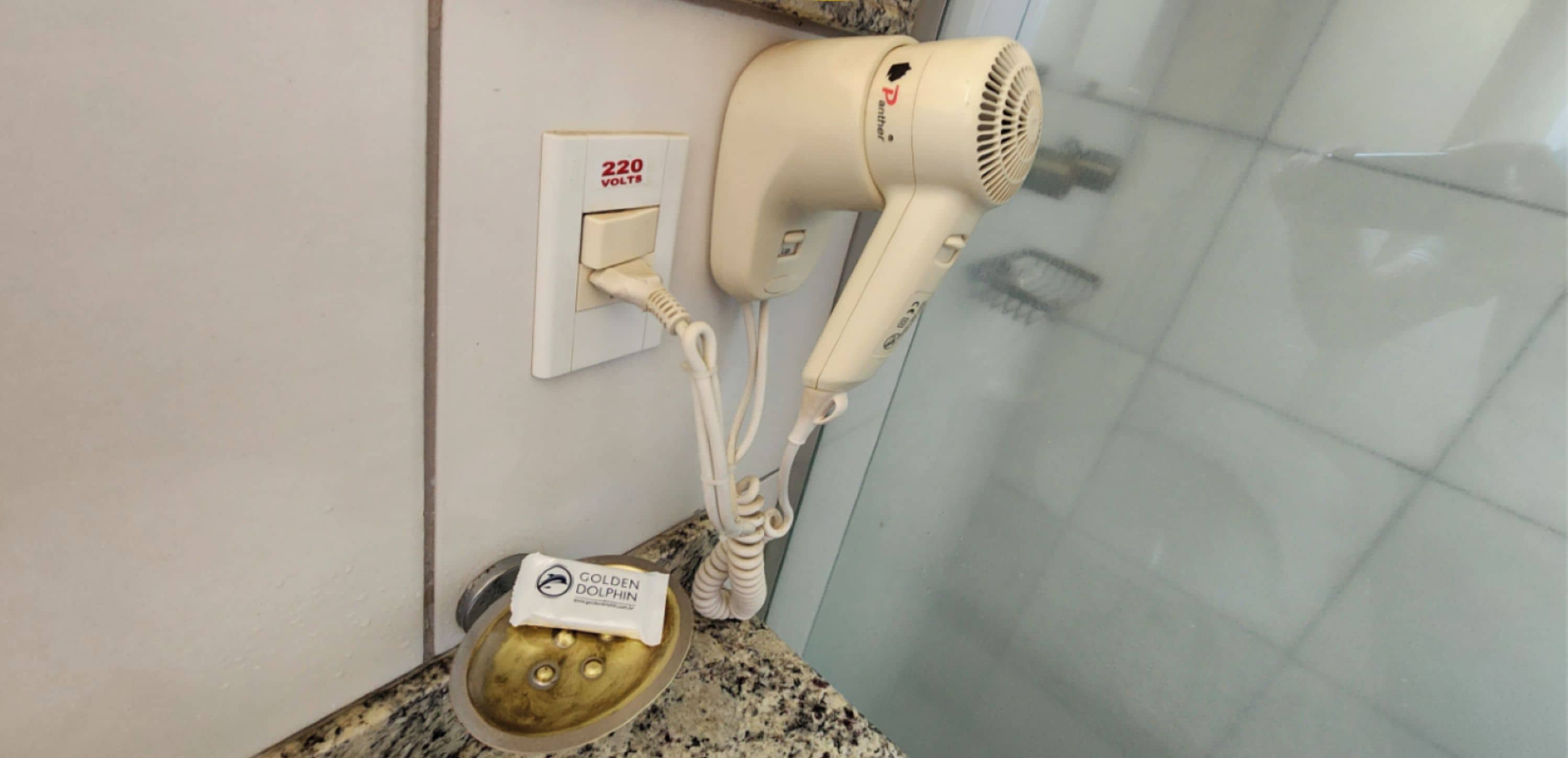 A hairdryer plugged into a wall socket with a 220 volts label, a water fountain, and a towel rack.