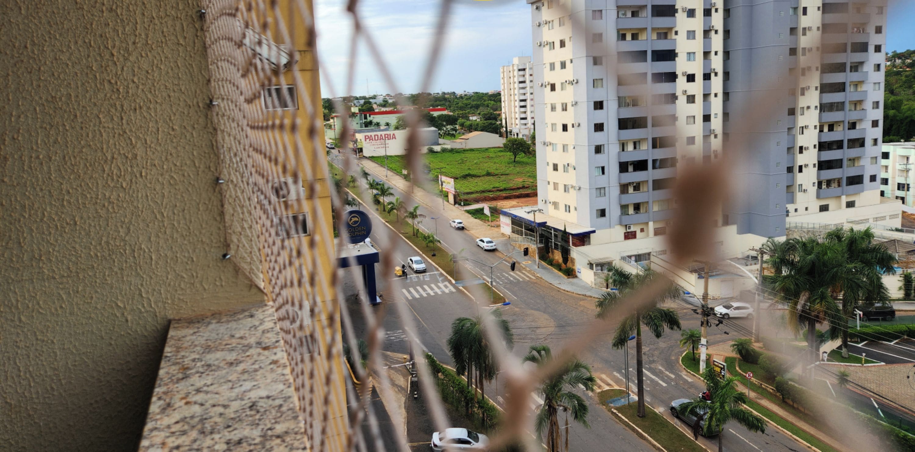 Um edifício alto com várias varandas, vista da rua com carros e uma placa indicando 'Padaria'.