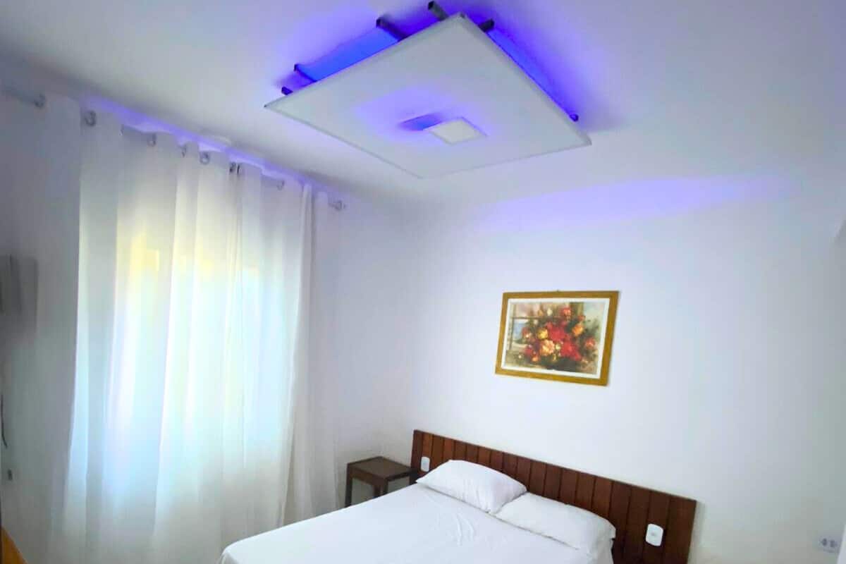 Habitación de hotel con una cama grande, una mesita de noche, una lámpara en el techo, un cuadro en la pared y una ventana con cortinas.