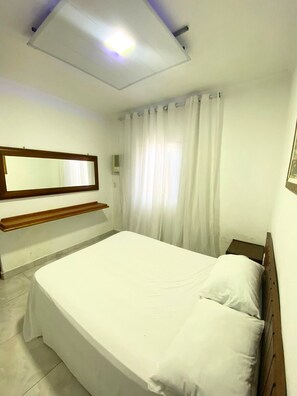 A neatly made bed in a room with a large mirror, a bedside table, and a window with curtains. - Pousada Ilha Bonita (Guarujá)