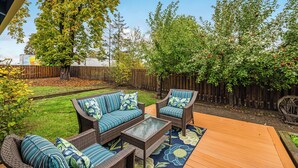 Ein Terrassenbereich im Freien mit Korbmöbeln und einem Couchtisch mit Glasplatte, umgeben von einer Holzterrasse und einem Rasen.
