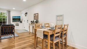 Ein Essbereich mit einem Holztisch, gedeckt für vier Personen, umgeben von Stühlen mit Kissen. Im Hintergrund ist eine Küchenzeile erkennbar, mit einem Fenster, das Grünpflanzen draußen zeigt.