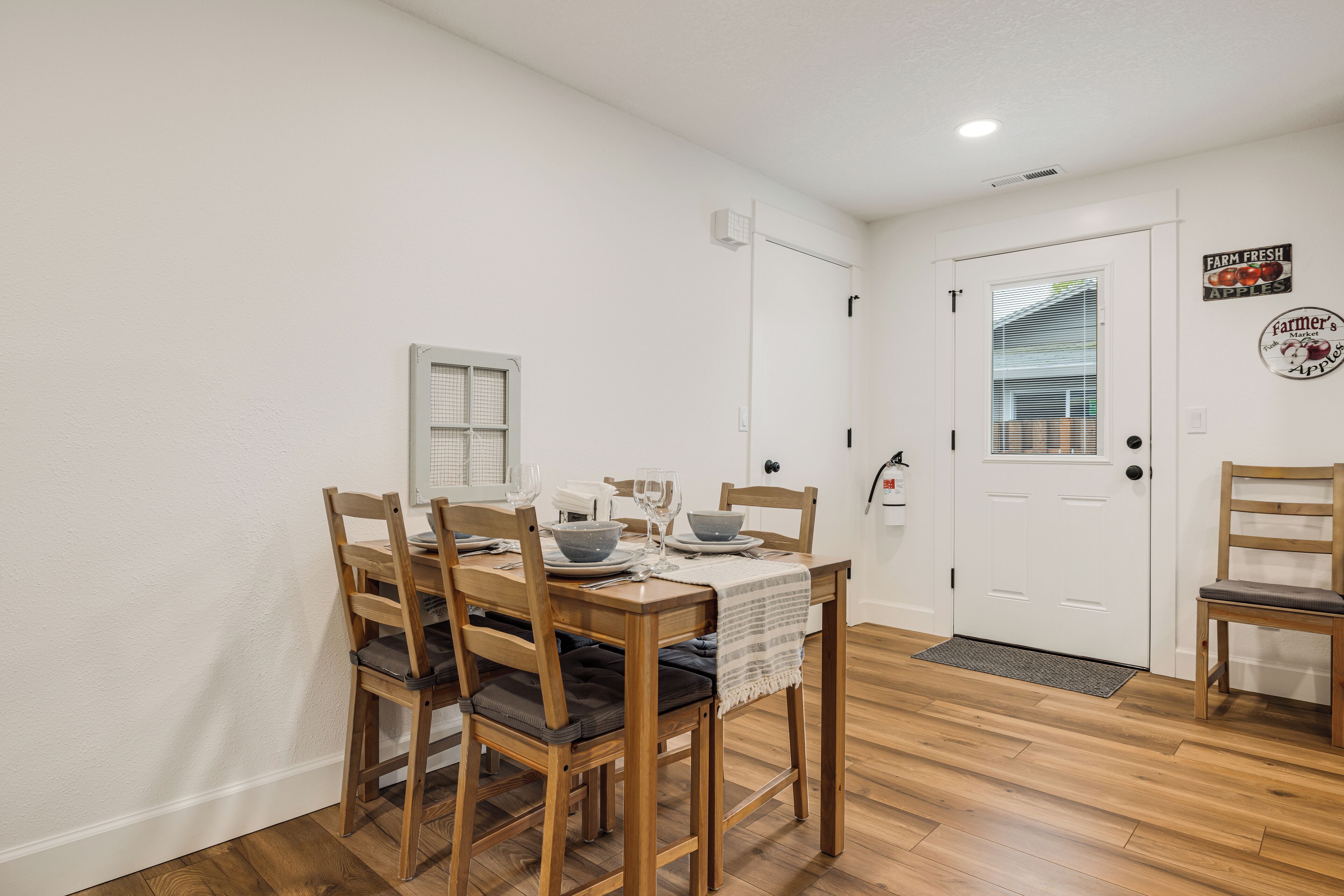 Un coin repas avec une table en bois dressée pour quatre personnes, entourée de chaises dotées de coussins. Une petite fenêtre avec des stores est visible sur le mur.