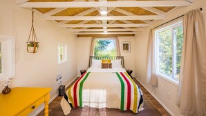 A bedroom with a wooden ceiling, a bed with a colorful striped blanket, a small bedside table, a nightstand, a window with curtains, and a hanging plant.
