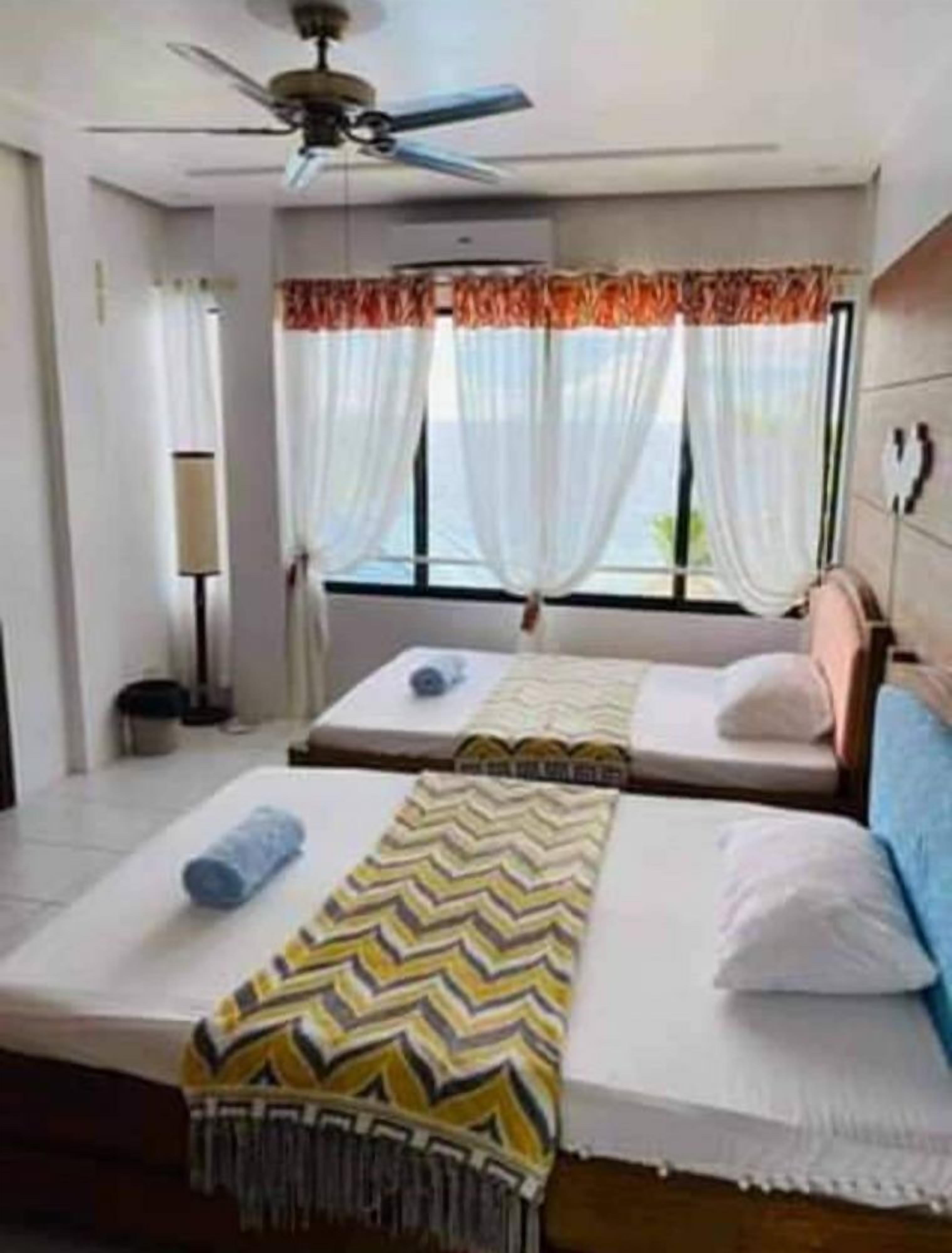 A hotel room with two beds, a ceiling fan, a window with curtains, and a bedside lamp.