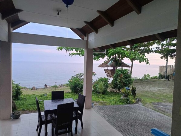 A covered patio with a table and chairs, overlooking a beach and ocean.