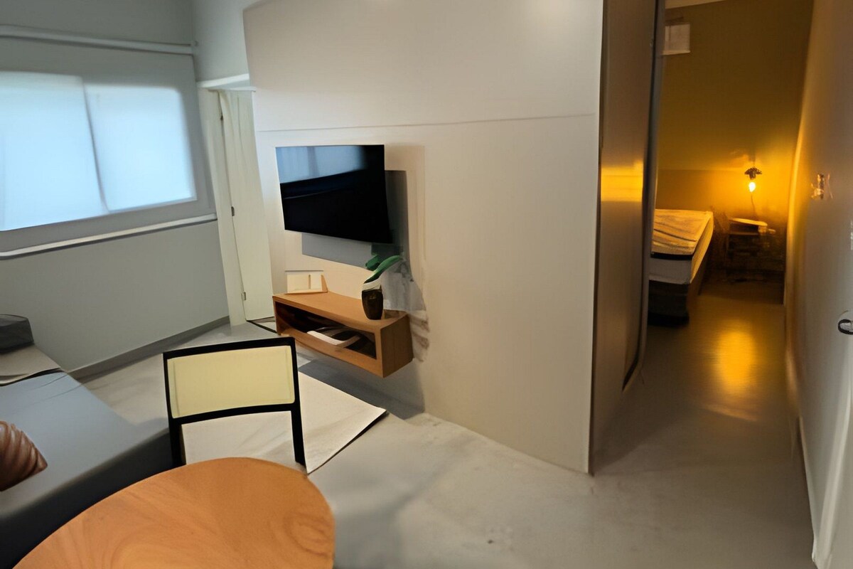A modern, compact living space with a flat-screen TV mounted on a wall, a small seating area with a table and chairs, and a narrow hallway leading to another room.
