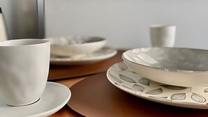Ruokapöydällä on valkoinen kuppi ja aluslautanen, valkoinen lautanen, jossa on lehtikuvio, ja valkoinen kulho.