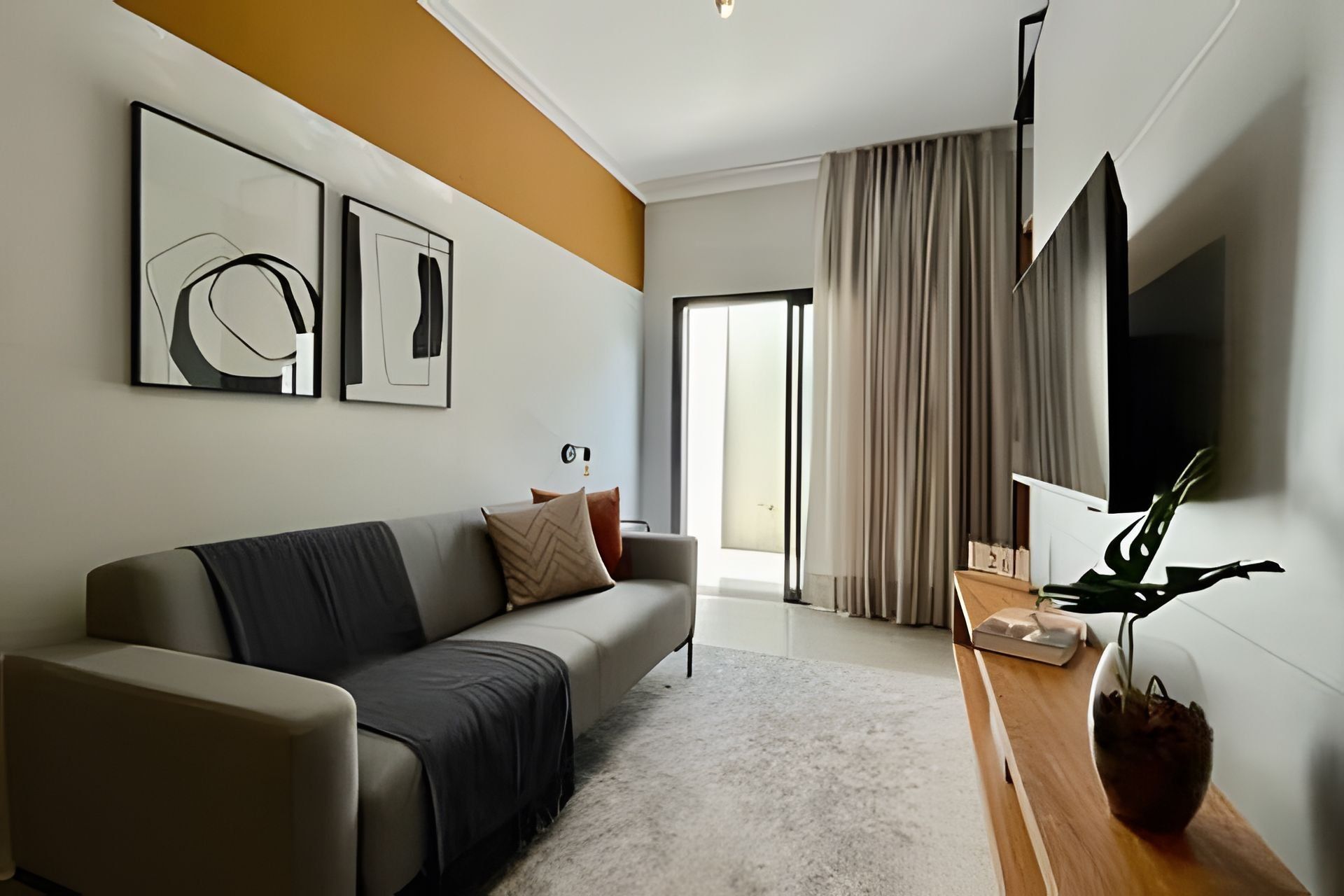 A modern living room with a sofa, a coffee table, and a flat-screen TV mounted on the wall. There is a large window with curtains, and artwork is displayed on the wall.