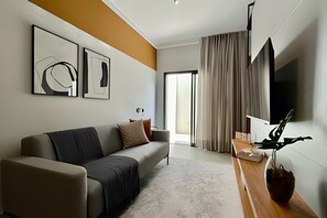 Ein modernes Wohnzimmer mit einer Couch, einem Couchtisch und einem an der Wand montierten Flachbildfernseher. Ein großes Fenster mit Vorhängen und Kunstwerke an der Wand.