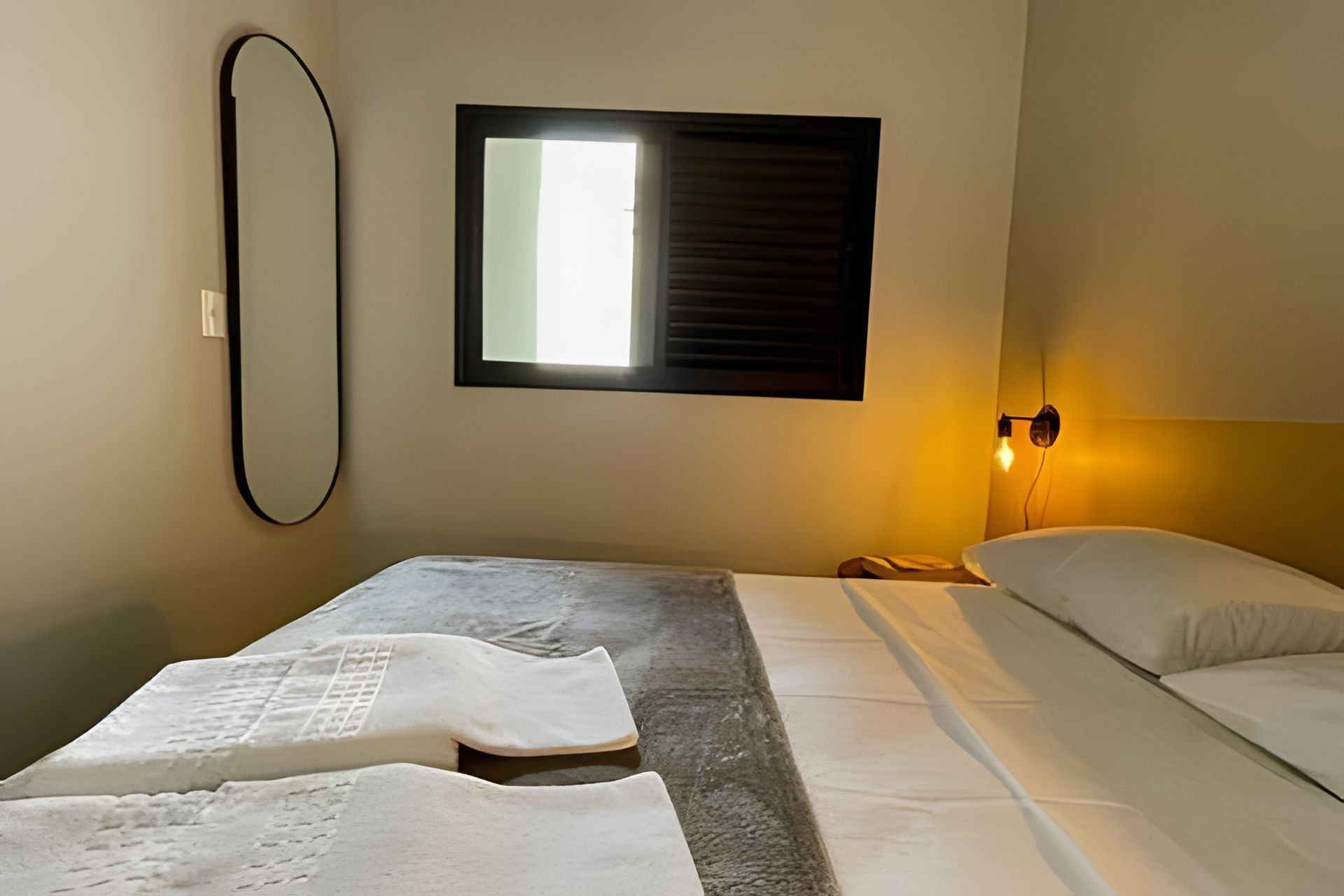 Quarto de hotel com cama, mesa de cabeceira, espelho fixado na parede e janela com persianas.