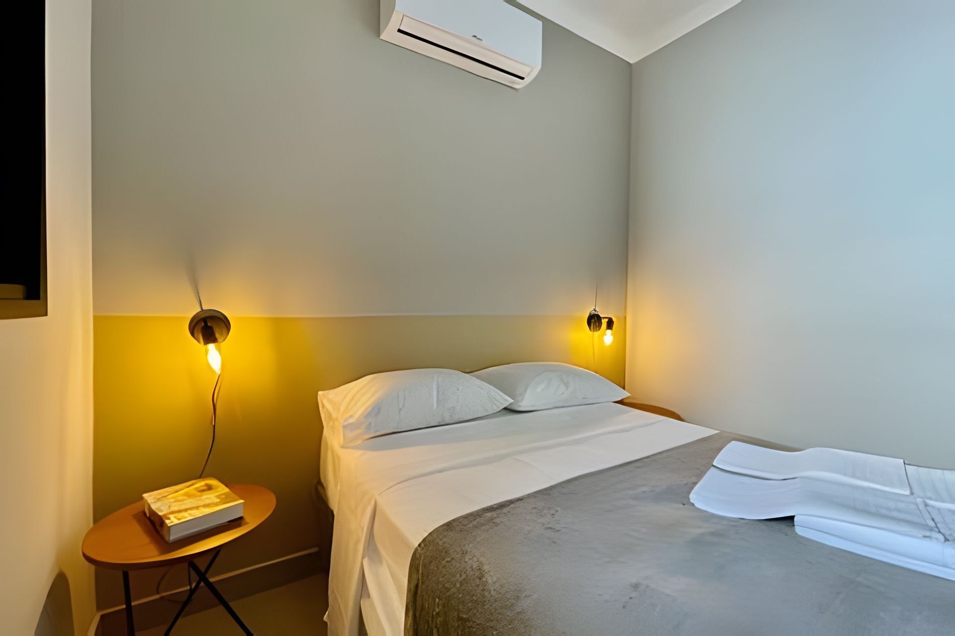 A hotel room with a bed, two wall-mounted reading lamps, a small round table with a book, and an air conditioning unit on the ceiling.