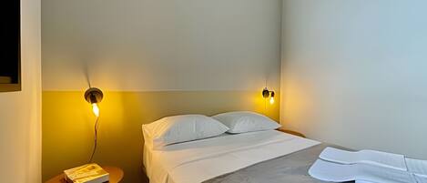Habitación de hotel con cama, dos lámparas de lectura fijadas a la pared, una mesita redonda con un libro y un equipo de aire acondicionado en el techo.