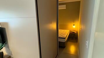 Corredor estreito que leva a um quarto com cama e mesa de cabeceira, uma luminária na parede e uma porta à direita.