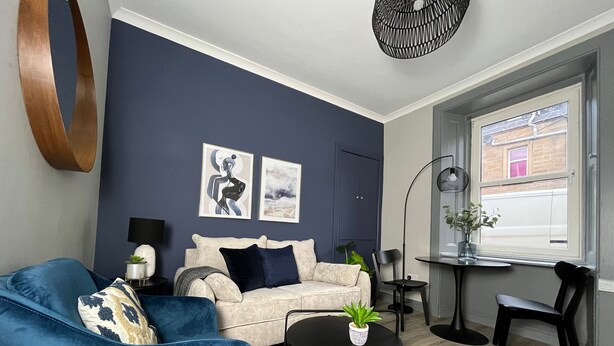 A modern living room with a blue sofa, a beige sofa, a round black coffee table, and a small side table. There are two framed artworks on the wall, a floor lamp, and a window with a view of a brick building. - Victoria Street (Galashiels)