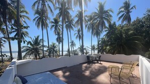 A balcony with a bed, a table, and chairs, overlooking a beach with palm trees and the ocean. - Sugarloaf Beachhouse and Surf Lodge (Jucuaran)