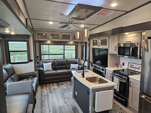 A compact RV interior with a kitchenette, a seating area with a sofa, and a large window providing a view. - RV14 | Coastal Retreat – Montana High Country (Bolivar Peninsula)