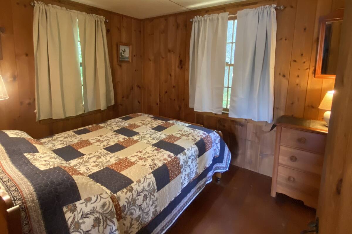 Quarto de cabana de madeira com uma cama, duas janelas com cortinas, um penteado e um abajur.