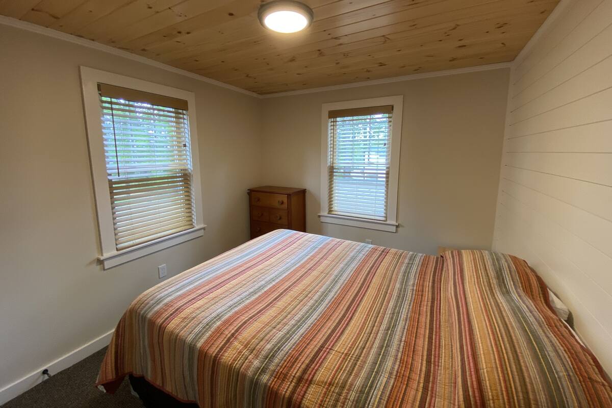 Quarto com teto de madeira, duas janelas com persianas, cama com colcha listrada e uma mesa de cabeceira.