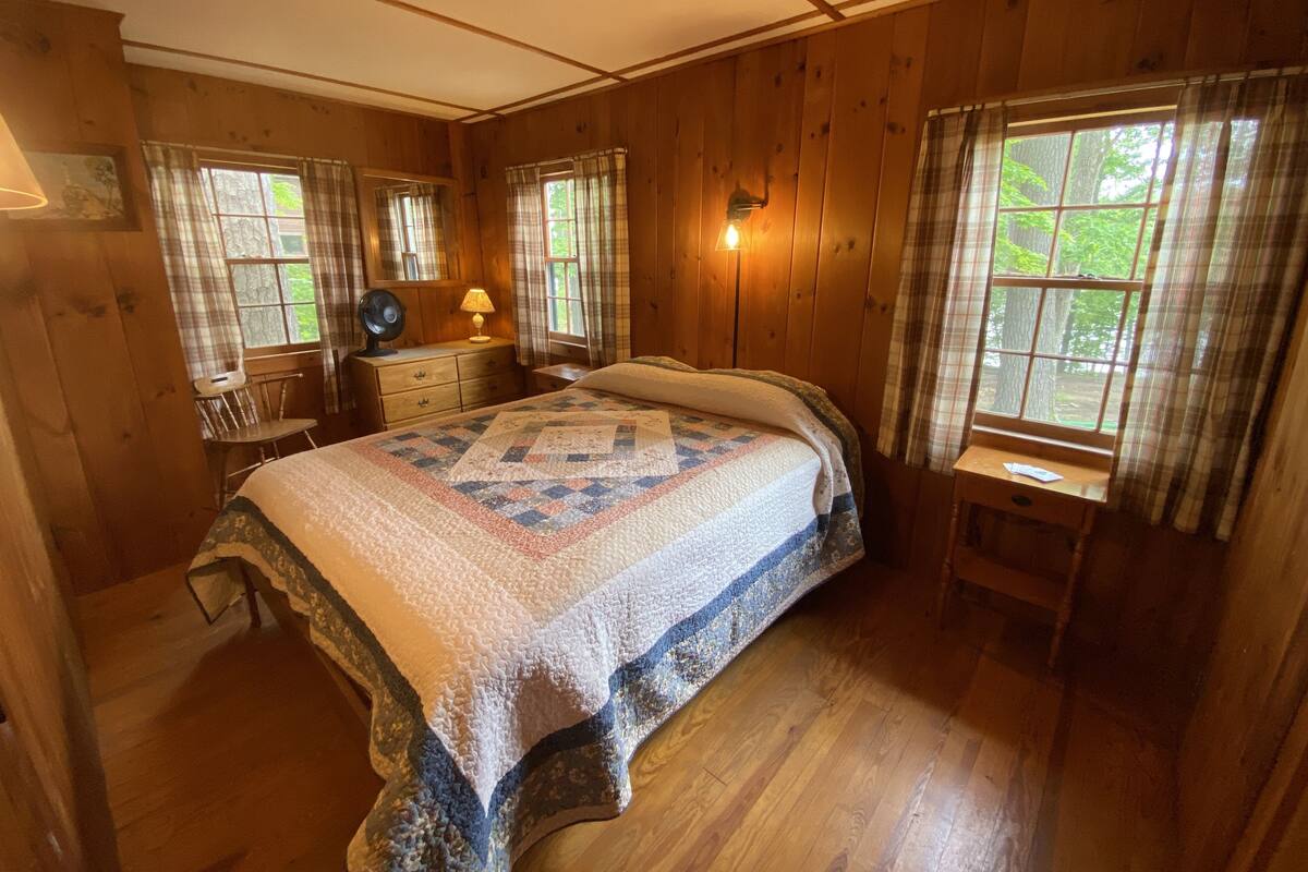 Quarto acolhedor de cabana de madeira com uma cama grande, uma mesa de cabeceira, cômoda e duas janelas com cortinas.