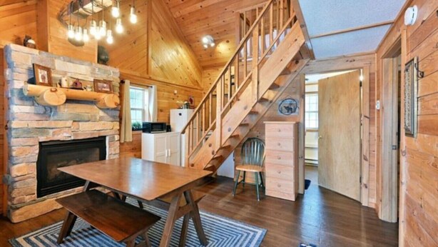 A rustic cabin interior with a stone fireplace, wooden dining table, and a staircase leading to an upper level. - Cozy Hayward Moose Lake Getaway (Hayward)