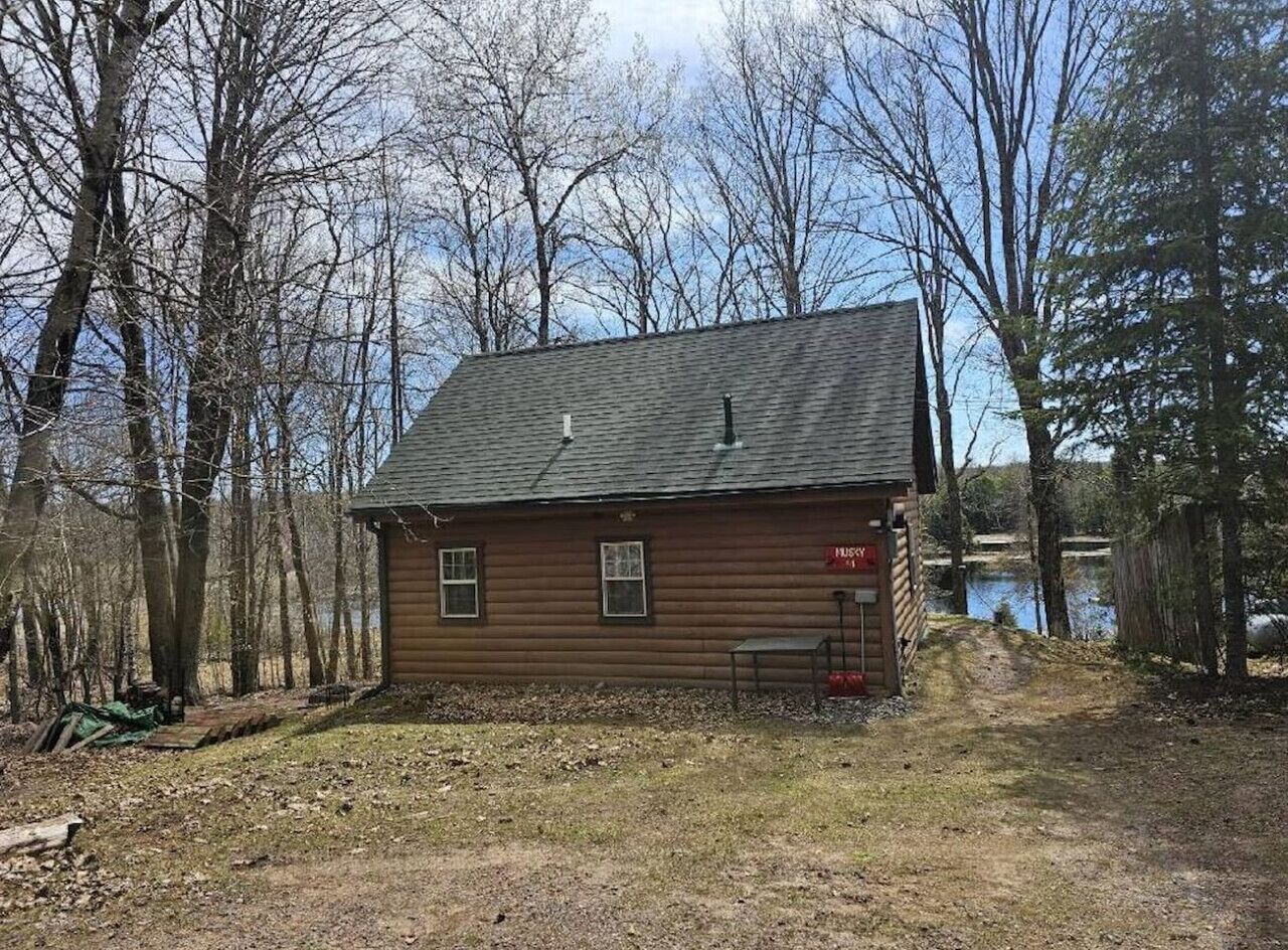 Une petite cabane en rondins, d’un seul étage, avec un toit en pente, située au bord d’un plan d’eau, elle-même entourée d’arbres.