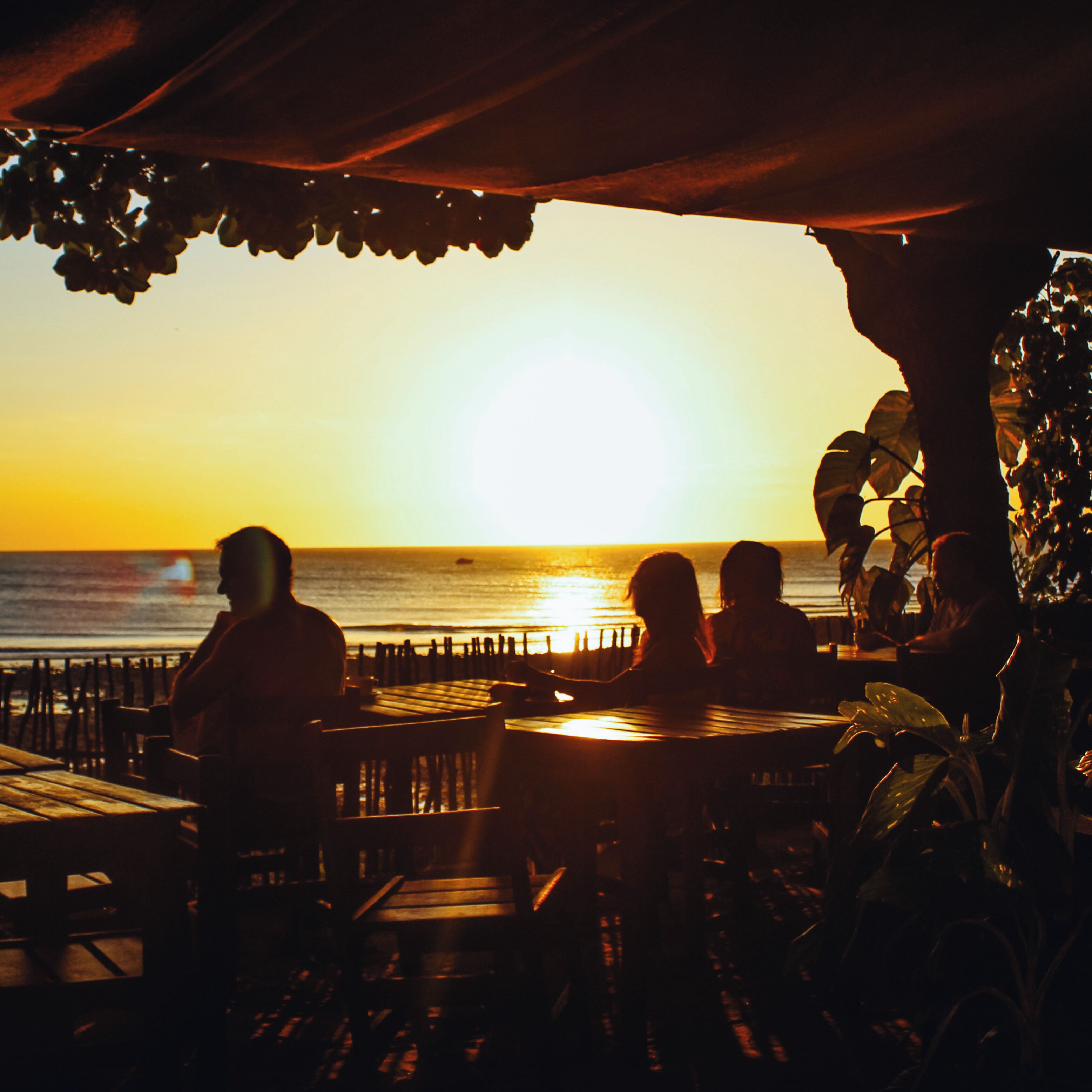A sunset view from an outdoor seating area with tables and chairs, overlooking the ocean.
