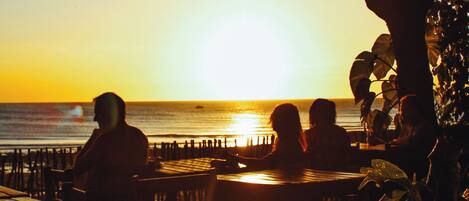 A sunset view from an outdoor seating area with tables and chairs, overlooking the ocean.