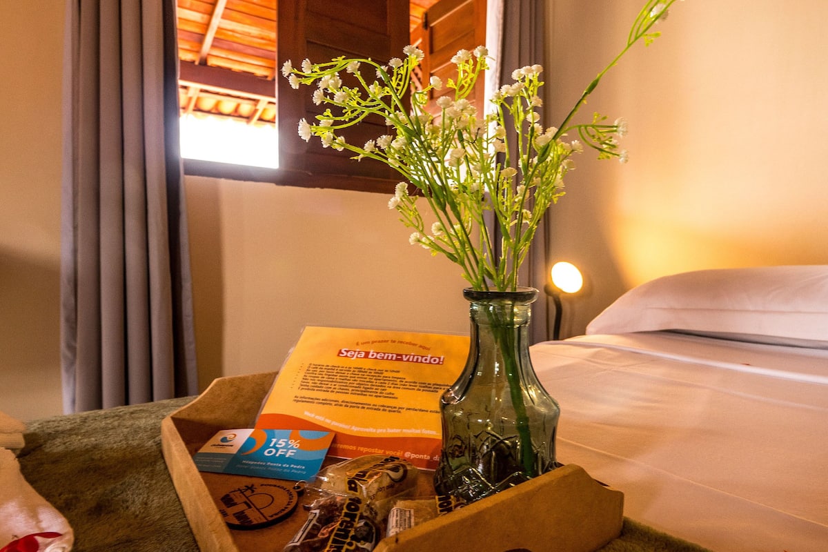 Une chambre d’hôtel avec un lit, une table de chevet ornée d’un vase de fleurs, un plateau avec une boîte et quelques objets, et une fenêtre avec des rideaux.