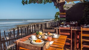 Une table en bois, dressée pour un repas, avec des boissons, donnant sur une plage où l’on aperçoit des vagues et un bateau au loin.