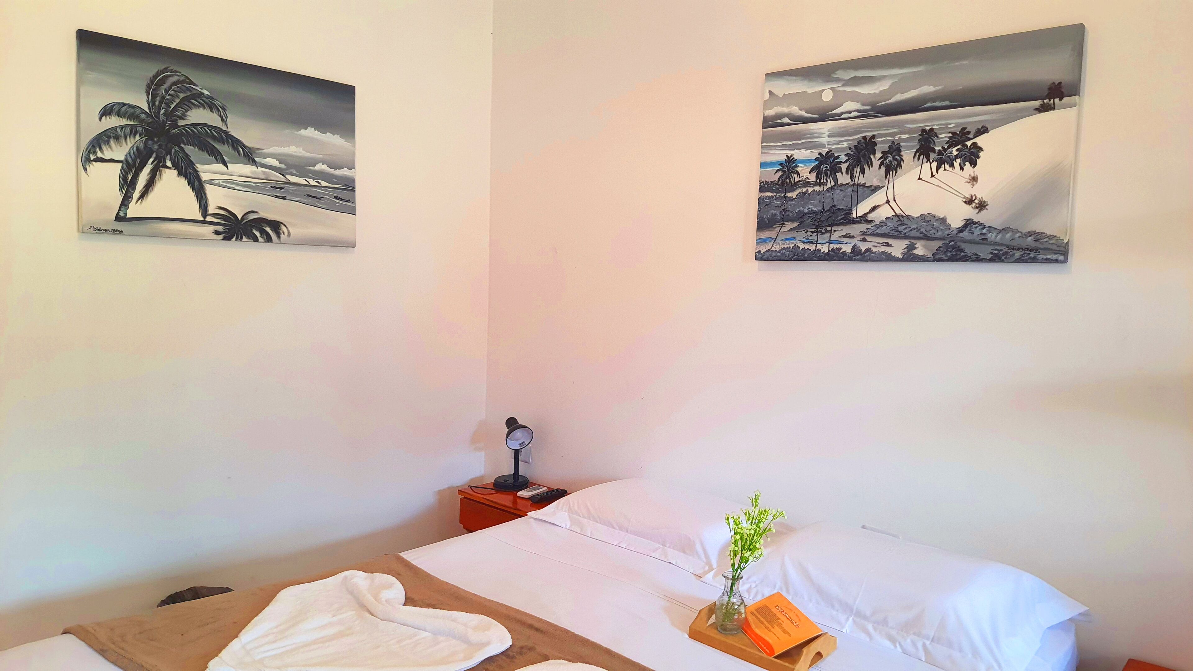 A hotel room with a bed, a nightstand, a lamp, a vase with flowers, and two wall-mounted paintings.