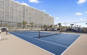 A tennis court with a net, surrounded by a fence, palm trees, and a large hotel building in the background. - Promo 6/27-7/1, Direct Beachfront & Beach Service, Perfect for Couples (Panama City Beach)